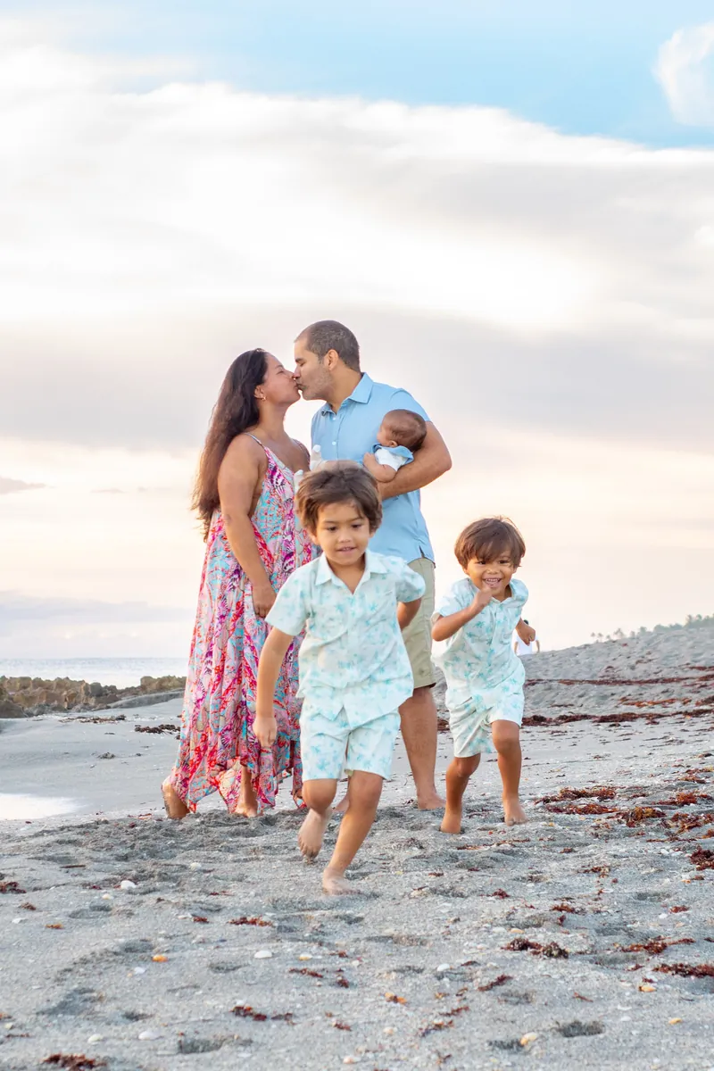 Parents sharing a kiss while holding their baby as two young boys run across the sand
