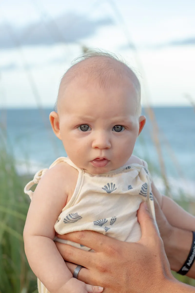 Baby with bright blue eyes held up beside the sea oats on a Jupiter beach